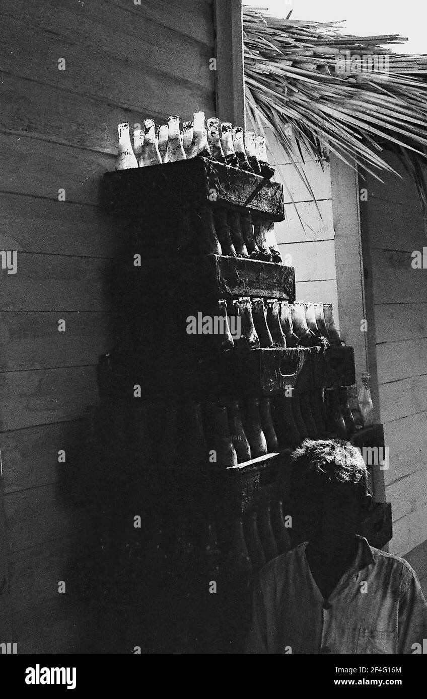 Une pile de bouteilles Coca Cola est visible sous un toit de chaume avec une personne en avant-plan dans les montagnes de la Sierra Maestra, Cuba, 1963. De la collection de photographies Deena Stryker. () Banque D'Images