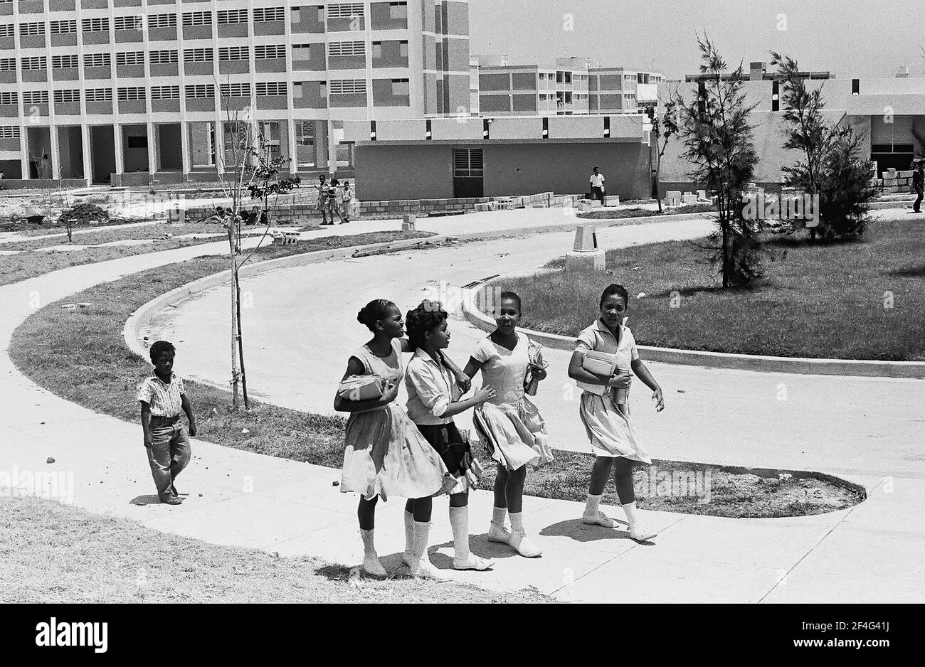 Le logement est visible à Habana Este, Cuba avec des enfants marchant dans un groupe sur un chemin près des bâtiments, un petit groupe devant un enfant plus jeune en retard, 1964. De la collection de photographies Deena Stryker. () Banque D'Images