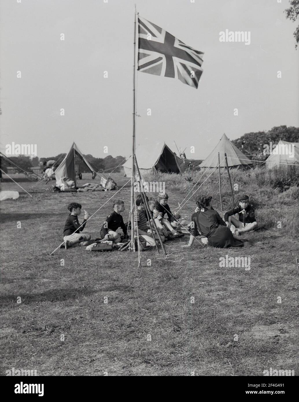 Camp scout des années 50, jeunes garçons assis à l'extérieur dans un champ avec un chef scout, une femme, autour d'un mât avec le drapeau Union Jack dans la zone de parc de Temple Newsam, Leeds, Angleterre, Royaume-Uni. Banque D'Images