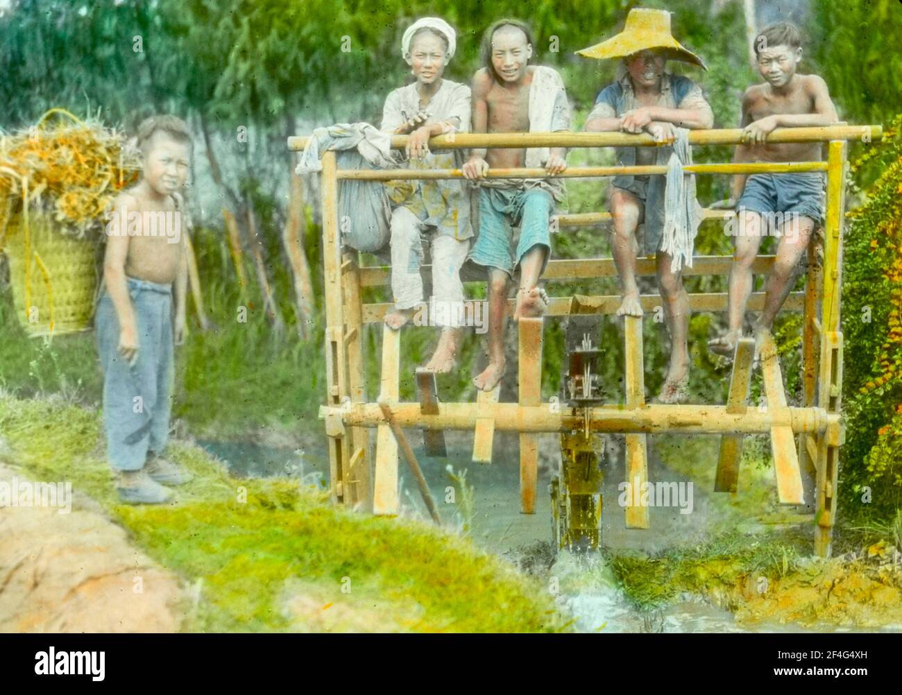 Plusieurs garçons utilisent leurs pieds pour pomper de l'eau à travers une roue en bois, avec un deuxième garçon portant un panier très chargé sur la rive de la rivière, Sichuan, Chine, 1917. De la collection de photographies Sidney D. Gamble. () Banque D'Images