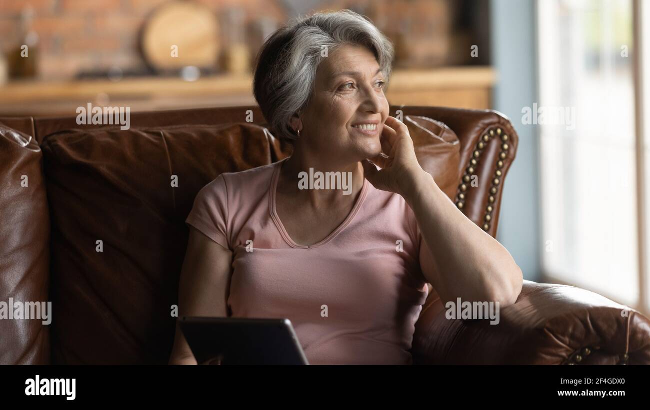 Une femme âgée s'assoit sur un canapé, rêve distrait de l'écran d'une tablette Banque D'Images