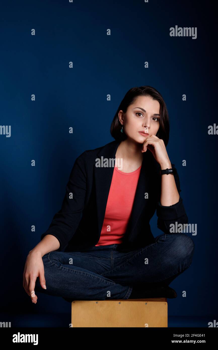 Portrait d'une jeune femme brune portant un débardeur corail, un blazer noir et un Jean bleu sur fond bleu foncé. Banque D'Images