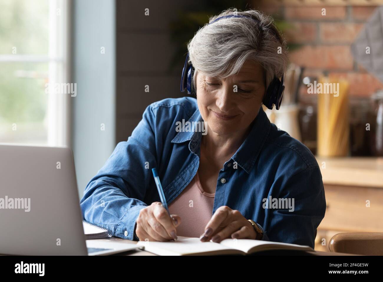 Femme âgée dans un casque regarder le séminaire en ligne sur ordinateur portable prendre des notes Banque D'Images