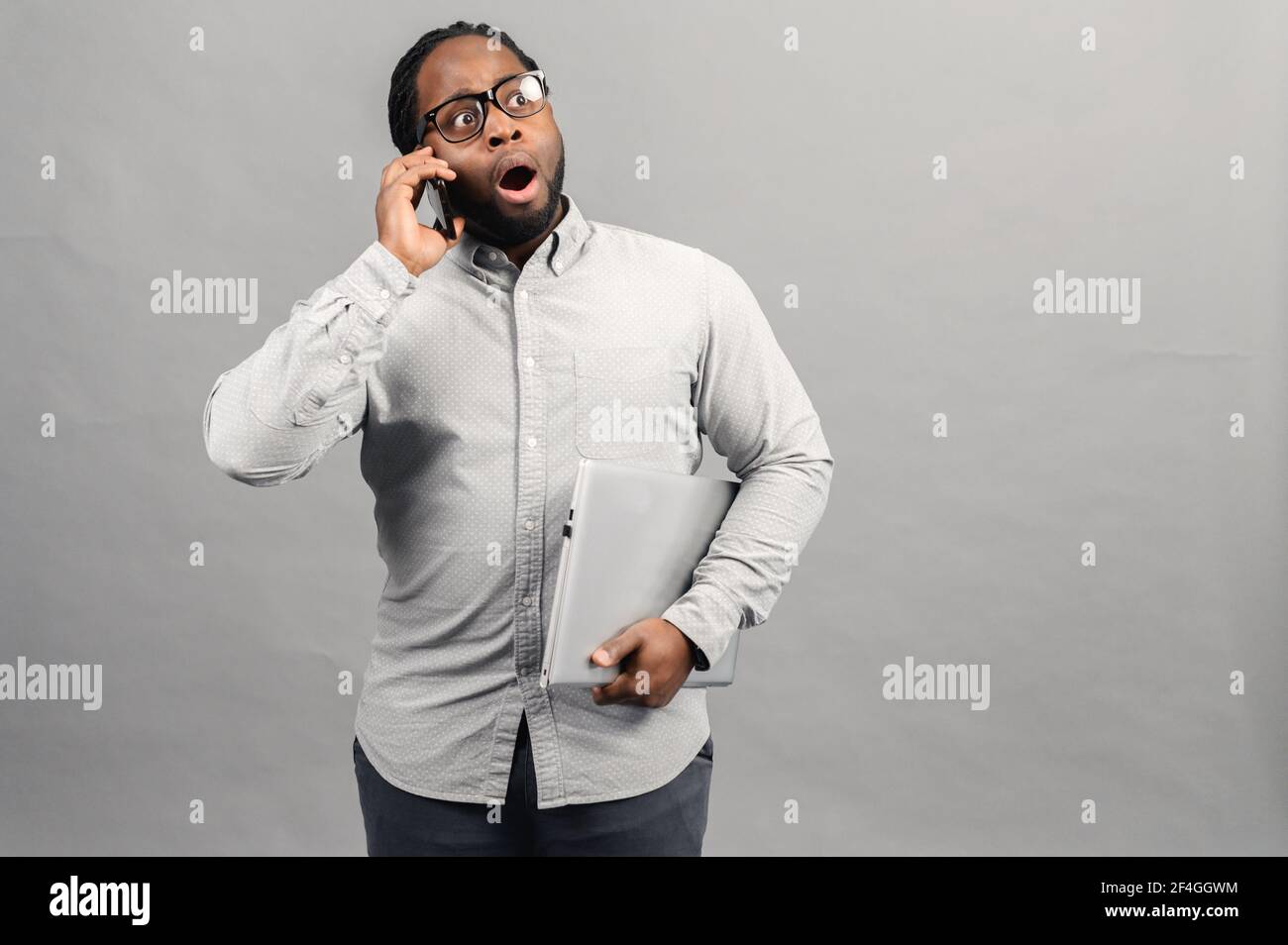 Jeune homme d'affaires latin parlant au téléphone sur fond gris isolé stressé avec un ordinateur portable dans la main, choqué par la honte et le visage surprise, en colère et frustré. Peur et contrarié par erreur. Banque D'Images