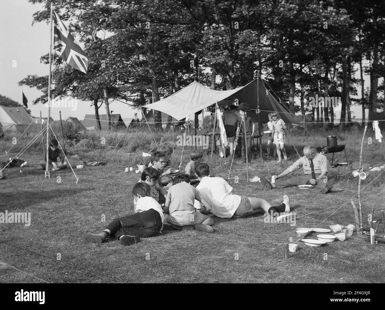 1969, historique, des garçons dans un camp de scouts d'été, assis sur l'herbe mangeant la nourriture, dans le domaine de Temple Newsam, Leeds, Angleterre, Royaume-Uni. Un drapeau de prise Union se trouve sur un mât. Banque D'Images