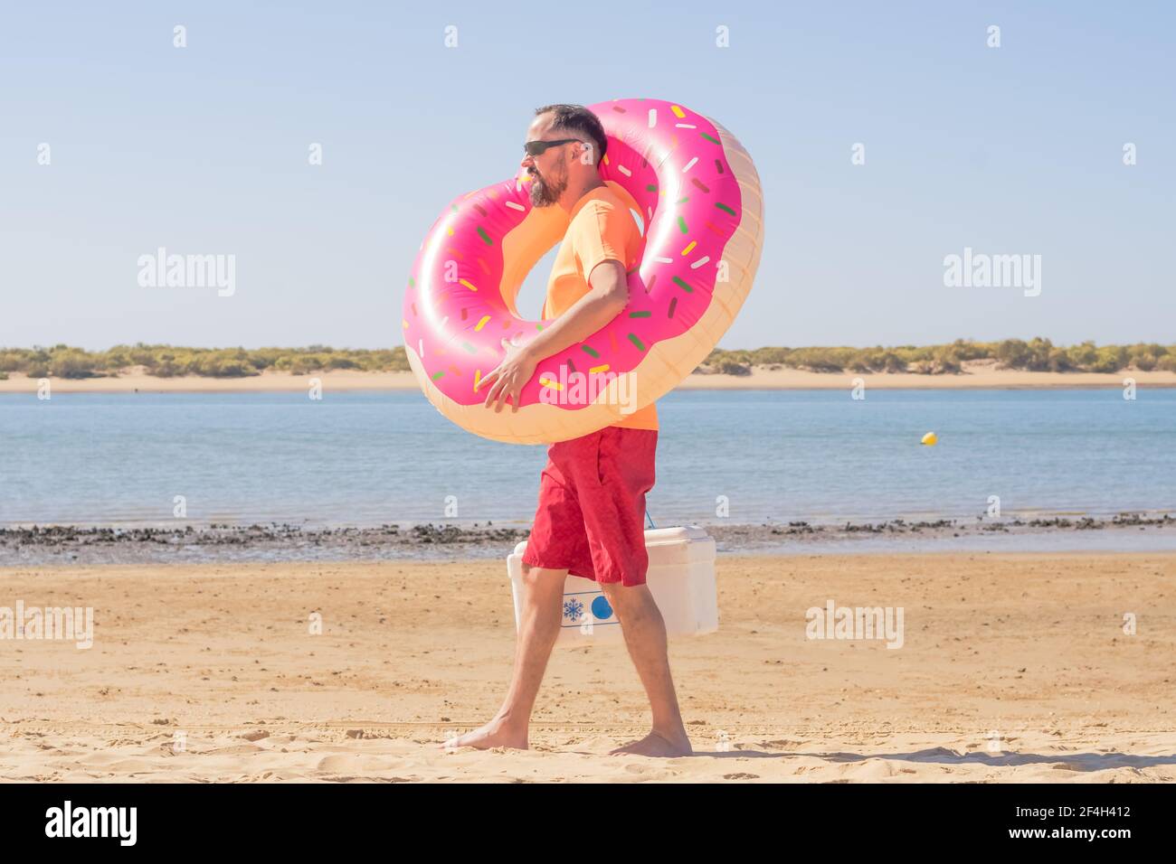 Un jeune homme hippster avec une barbe, des caleçons de natation et des lunettes de soleil porte un flotteur gonflable rose et un réfrigérateur sur la plage. Loisirs par une journée ensoleillée sur va Banque D'Images