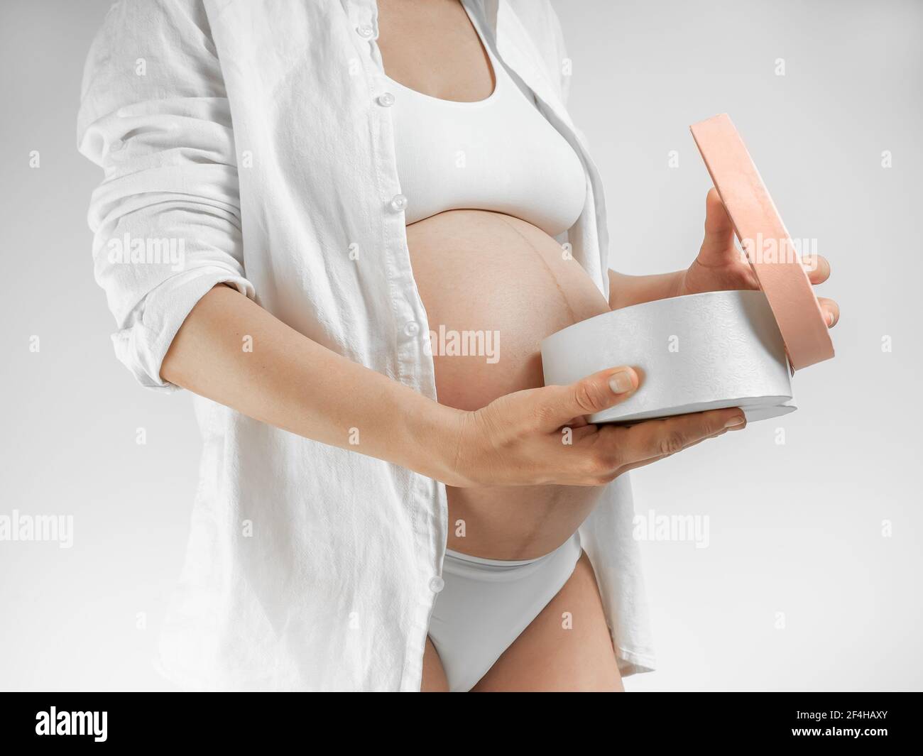 Dame dans le troisième trimestre de la grossesse. Une jeune femme enceinte en chemise blanche ouvre une boîte cadeau sur fond blanc. Une fille dans le dernier mois de prégnane Banque D'Images