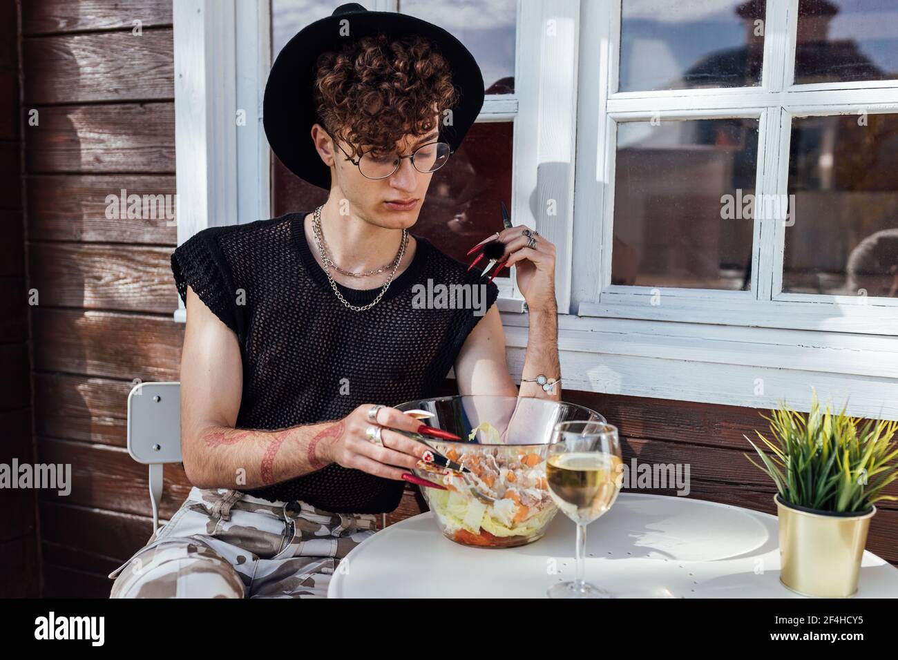 Jeune gay dans une tenue tendance assis avec des jambes croisées à table avec nourriture contre bungalow Banque D'Images