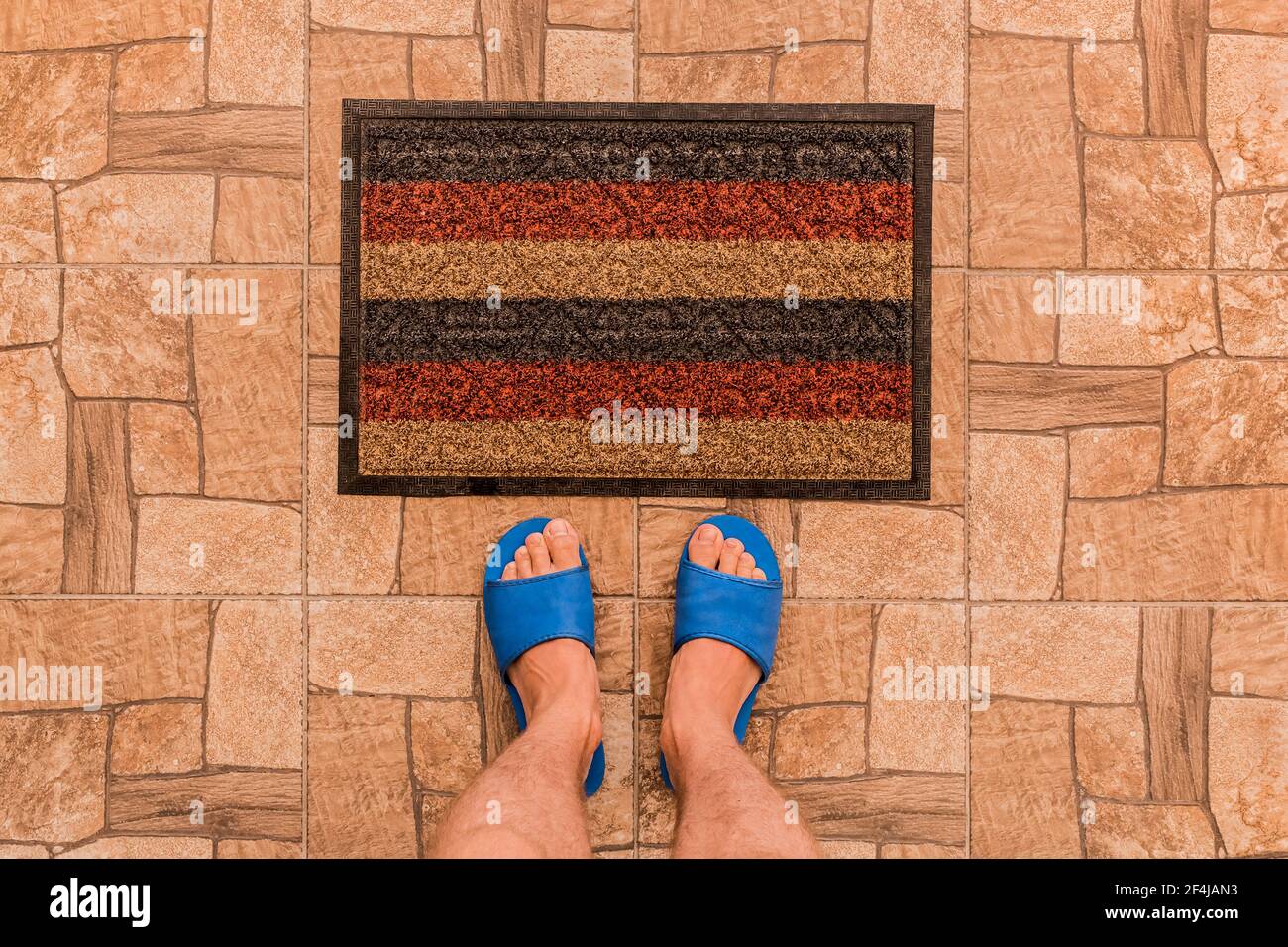 Les pieds mâles dans des pantoufles bleues se tiennent devant un tapis de pied sur un fond de sol carrelé marron texturé, vue du dessus. Banque D'Images
