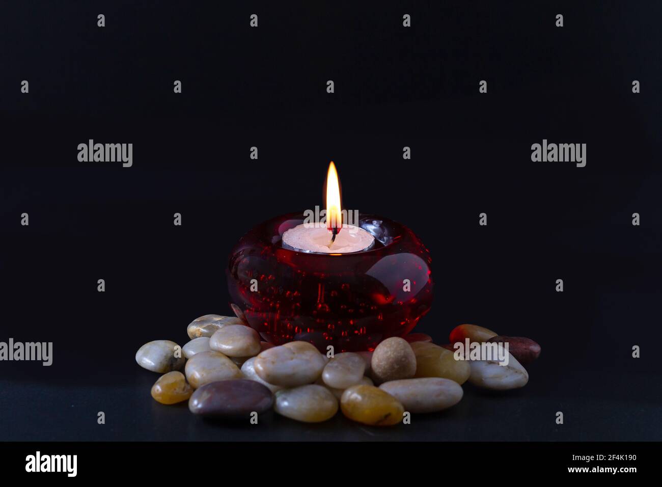Concept de deuil ou de prière. Bougie brûlante dans un chandelier rond rouge avec des pierres de couleur qui l'entourent. Tout cela sur fond noir. Banque D'Images