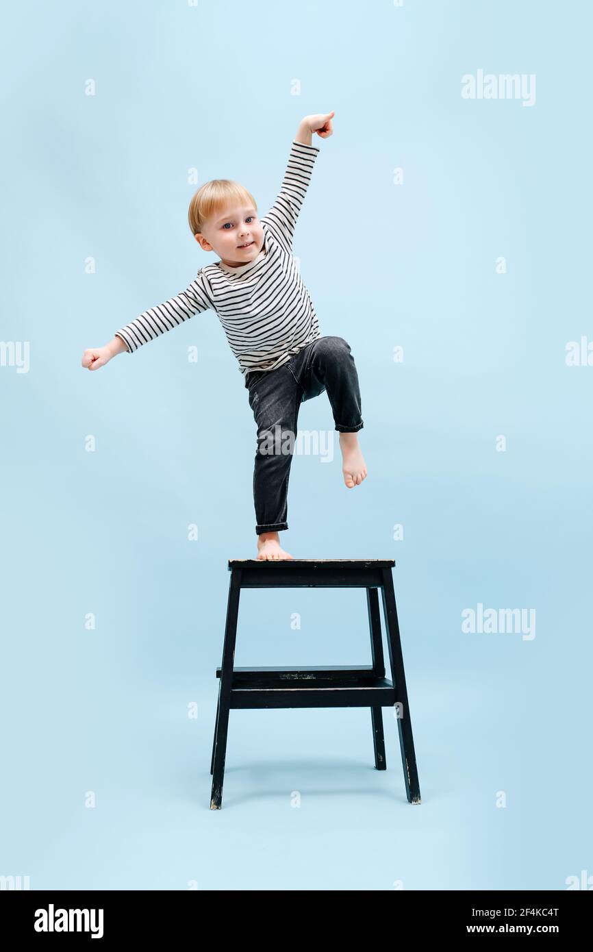 Agile pieds nus blond garçon en équilibre sur un pied sur un tabouret de  marche, inclinaison sur le côté. Il porte une chemise rayée à manches  longues. Prise de vue en studio.