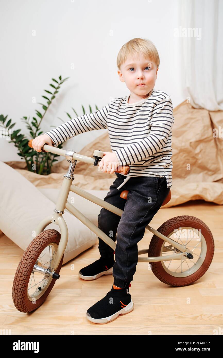 Beau petit garçon blond assis sur un petit vélo beige à deux roues à l'intérieur d'une chambre. Il porte une chemise rayée à manches longues. Papier d'emballage en arrière-plan. Banque D'Images