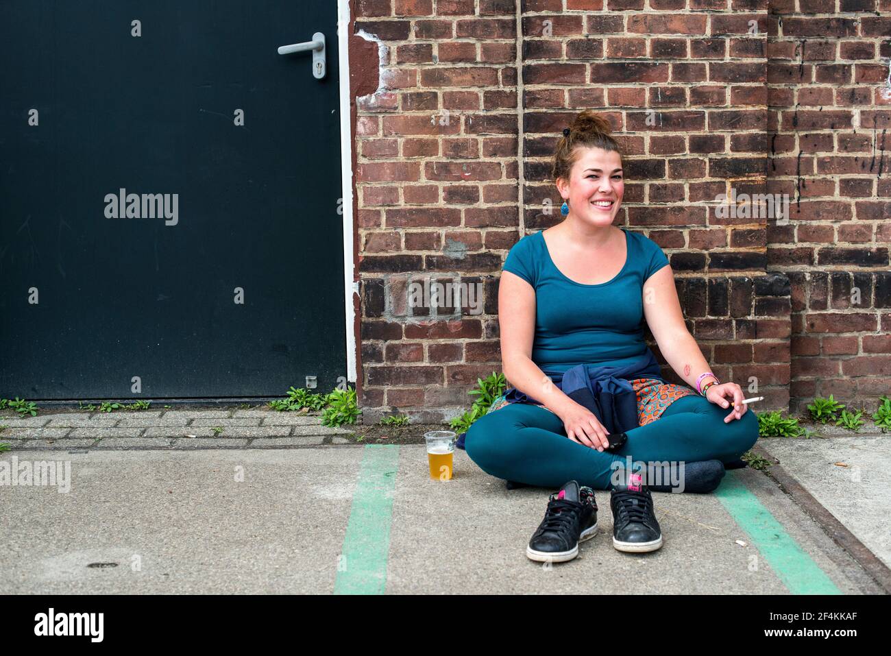 Tilburg, pays-Bas. Jeune, femme visiteur du Festival Mundial 2016 assis contre un vieux mur tout en appréciant une cigarette et un verre de bière. Banque D'Images