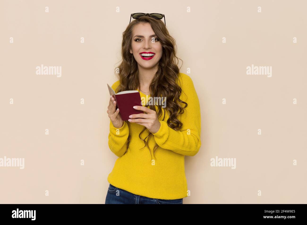 Une jeune femme heureuse en chandail jaune tient un passeport rouge ouvert, regarde la caméra et parle. Taille haute, prise en studio sur fond beige. Banque D'Images
