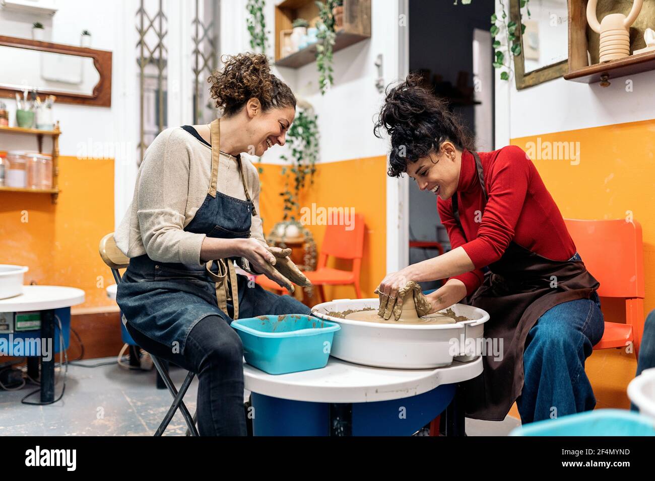 Photo de stock de femmes heureuses en tablier travaillant derrière une roue de potier dans un atelier. Banque D'Images