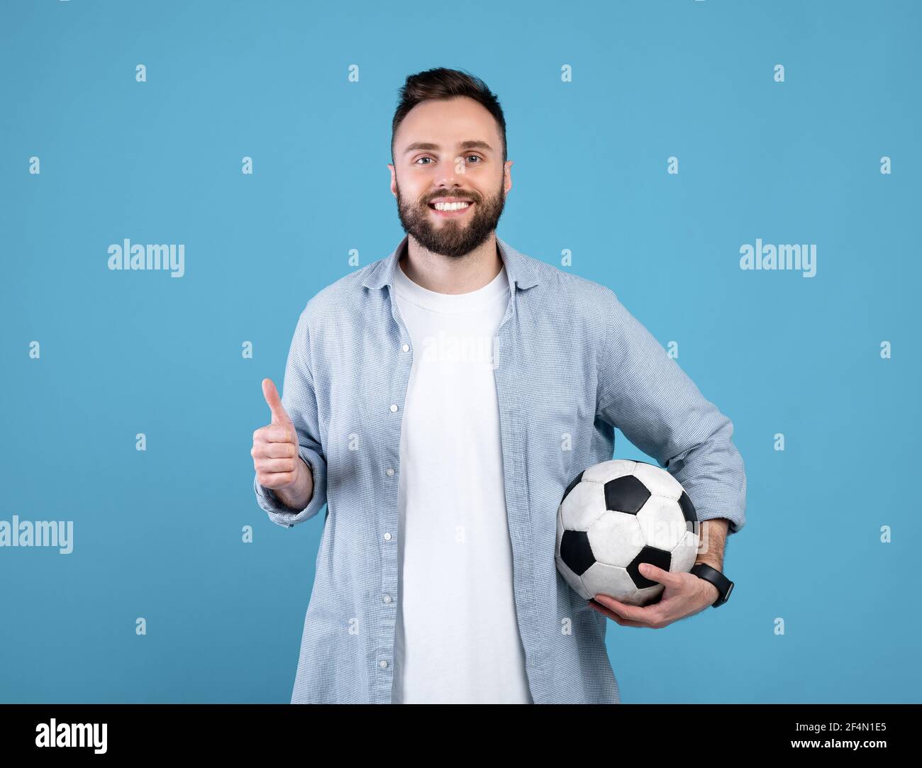 Jeune homme joyeux tenant le ballon de football, faisant le pouce vers le  haut de geste, recommandant le sport sur fond bleu studio Photo Stock -  Alamy