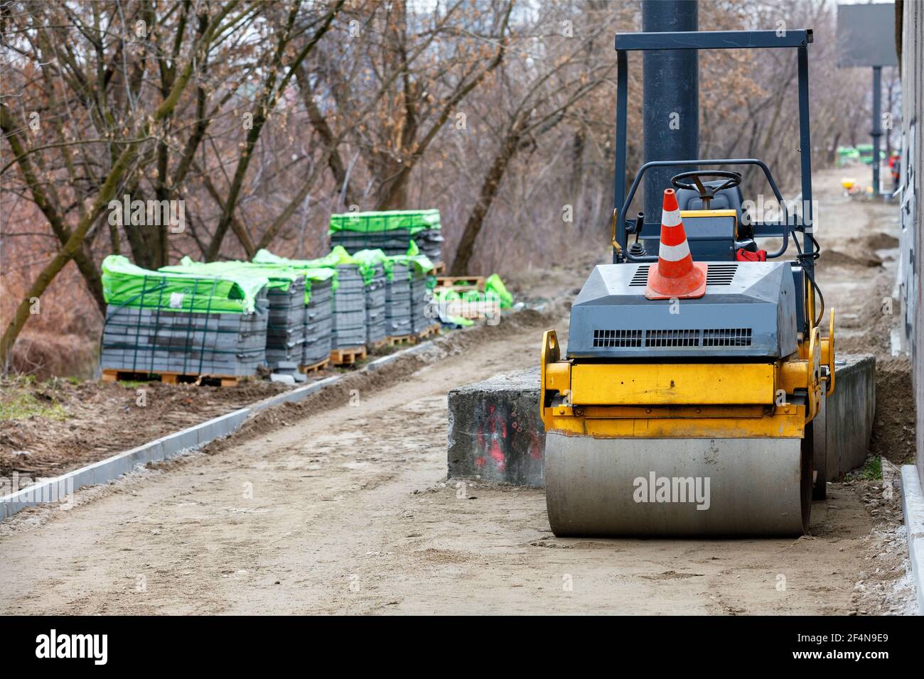 Un rouleau de route sur un chantier clôturé avec un trottoir en construction à l'arrière-plan et des piles de pavés gris en flou. Banque D'Images