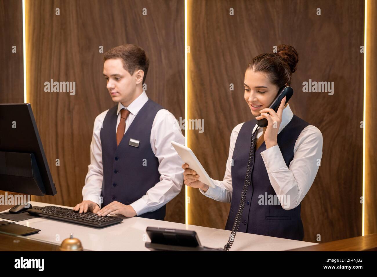 L'un des deux jeunes réceptionnistes de l'hôtel debout au comptoir, regardant l'écran tactile et consultant le client au téléphone contre un collègue Banque D'Images