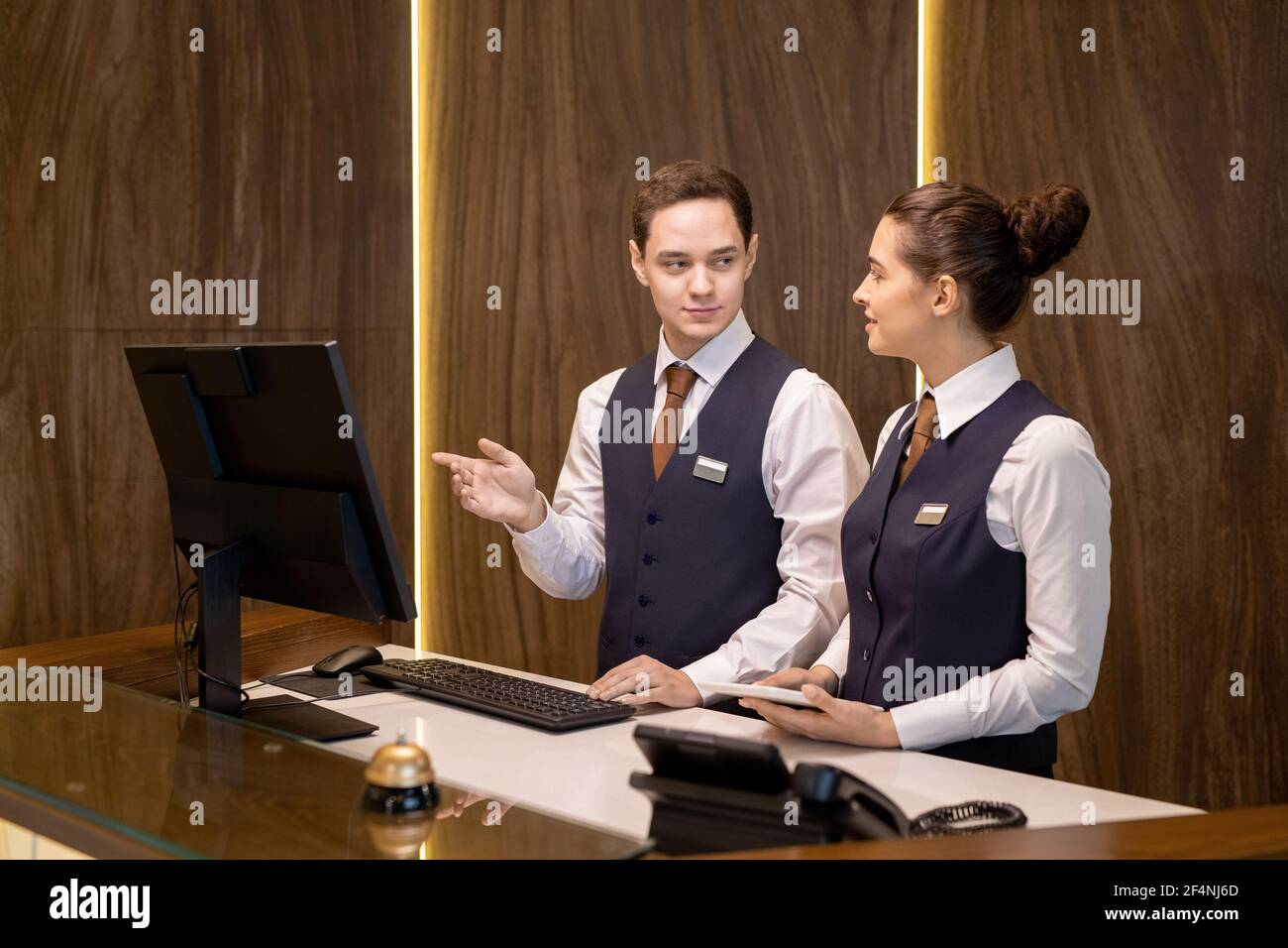 L'un des deux jeunes réceptionnistes de l'hôtel se tenant au comptoir, regardant un collègue avec pavé tactile et pointant vers l'écran d'ordinateur Banque D'Images