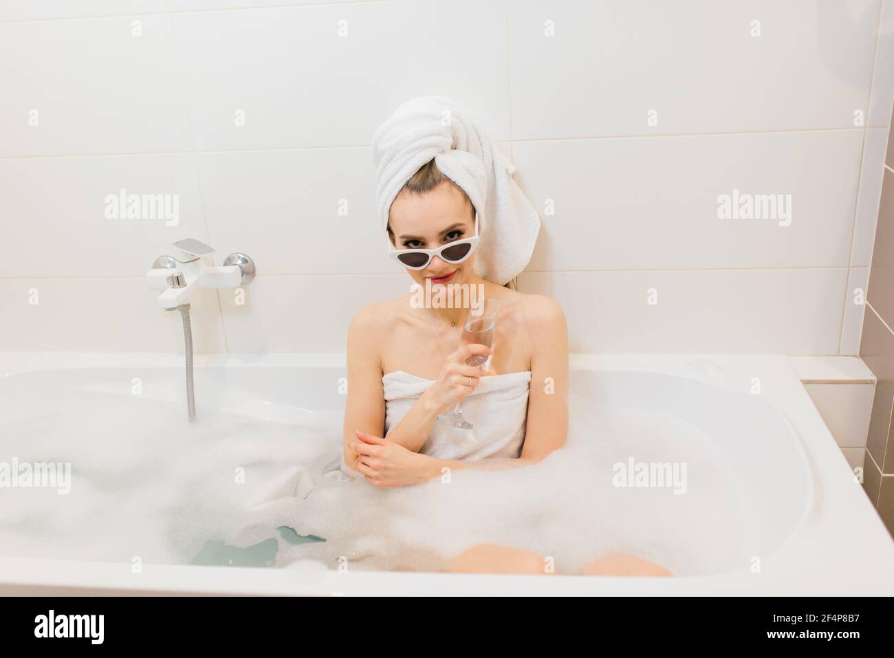 Jolie femme avec un sourire blanc neige dans la salle de bains. Femme élégante dans une serviette sur la tête et lunettes de soleil prend des soins spa à la maison ou à l'hôtel Banque D'Images