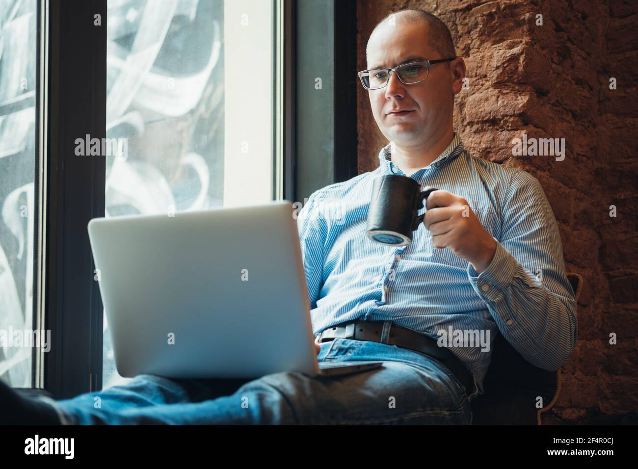 Homme dans une chemise bleue travaillant à la maison à l'aide d'un ordinateur portable près de la fenêtre. Travail à distance, travail en ligne, travail à domicile. Banque D'Images