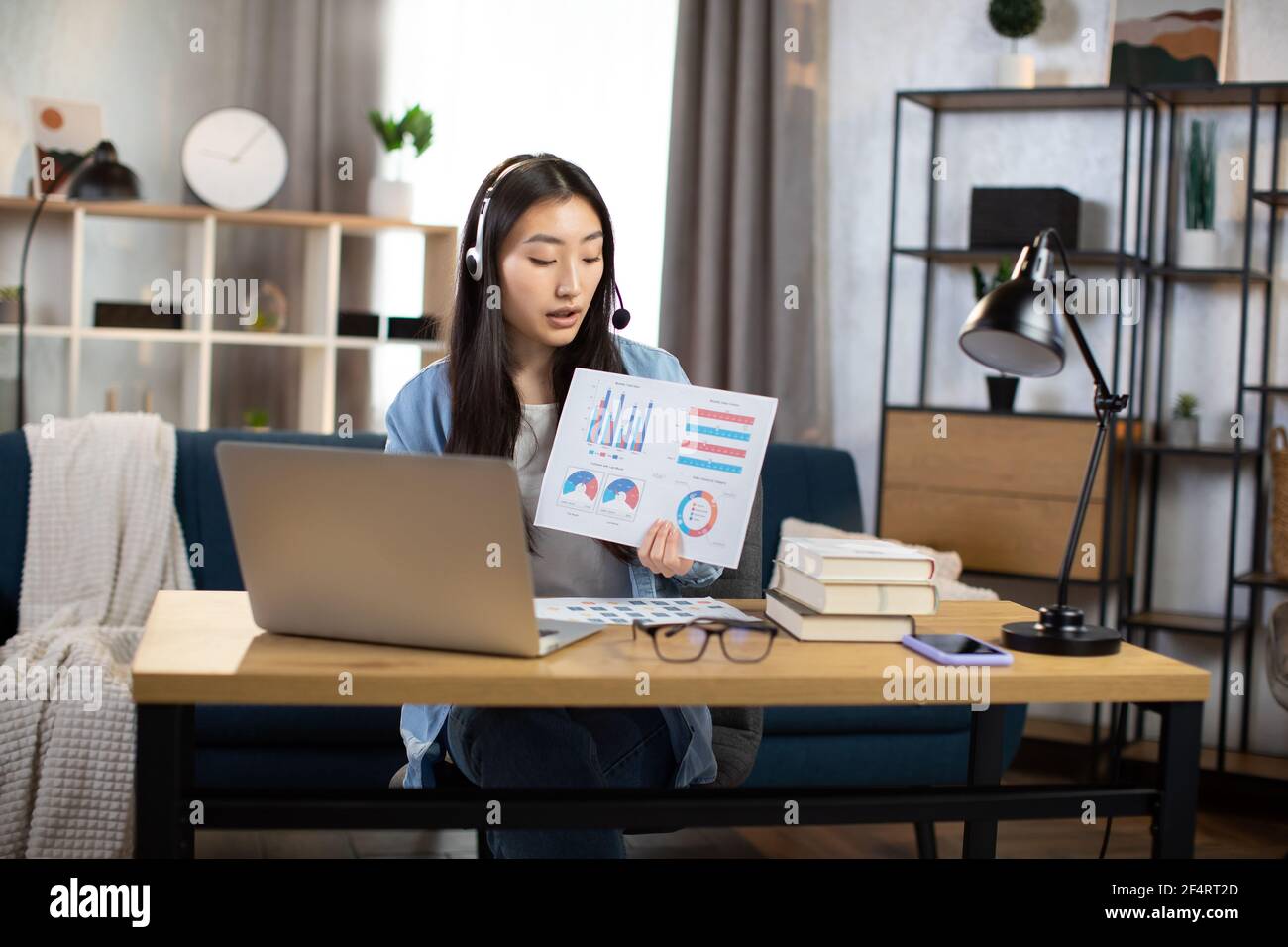 Jeune femme coréenne professionnelle présentant des graphiques et des graphiques financiers à ses collègues par vidéoconférence sur ordinateur portable. Femme indépendante ou responsable assis sur le lieu de travail et parlant dans un micro-casque. Banque D'Images