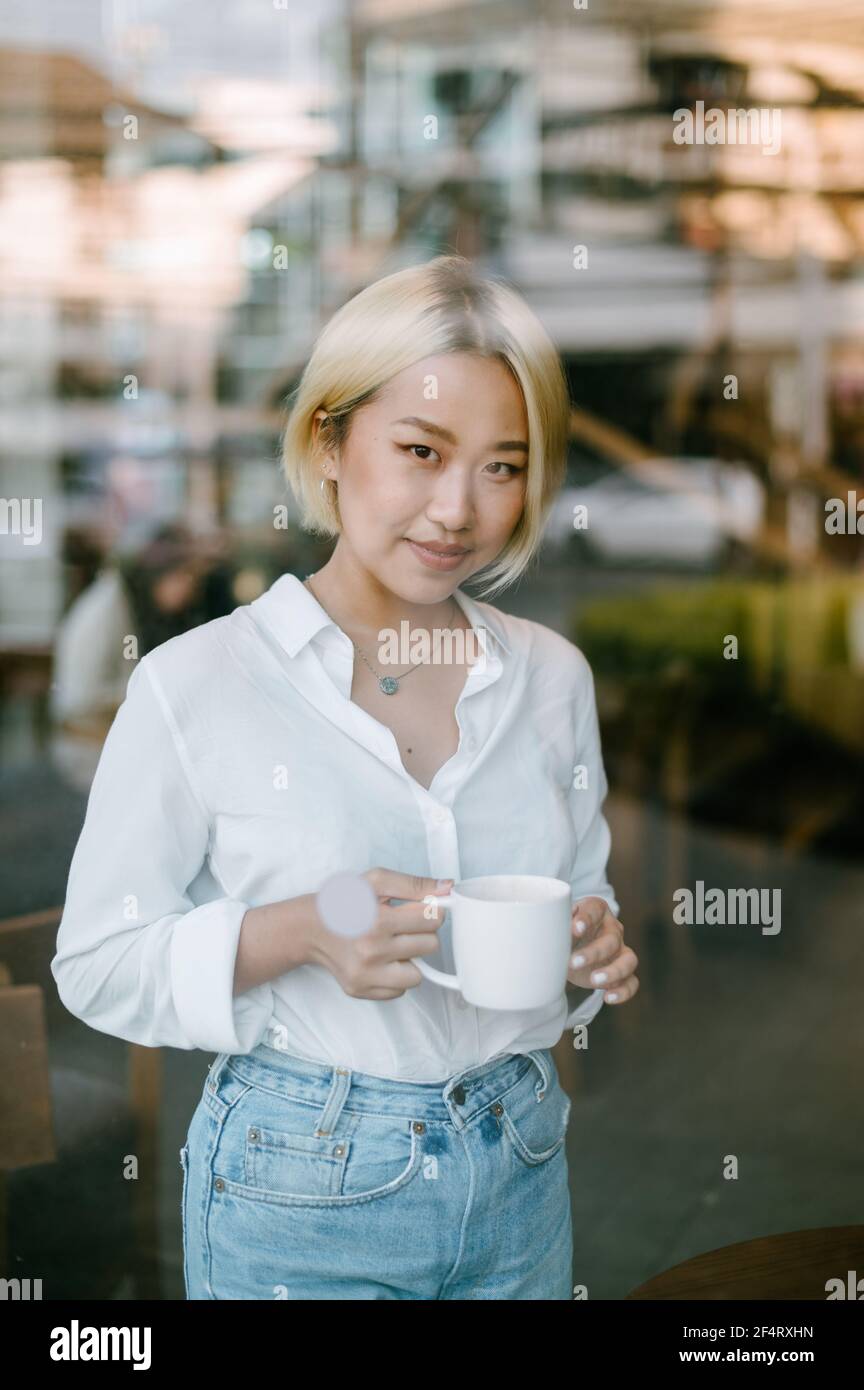 Une jeune femme asiatique avec une tasse de café dans ses mains est debout à l'intérieur derrière une grande fenêtre, regardant la caméra à l'extérieur. Banque D'Images