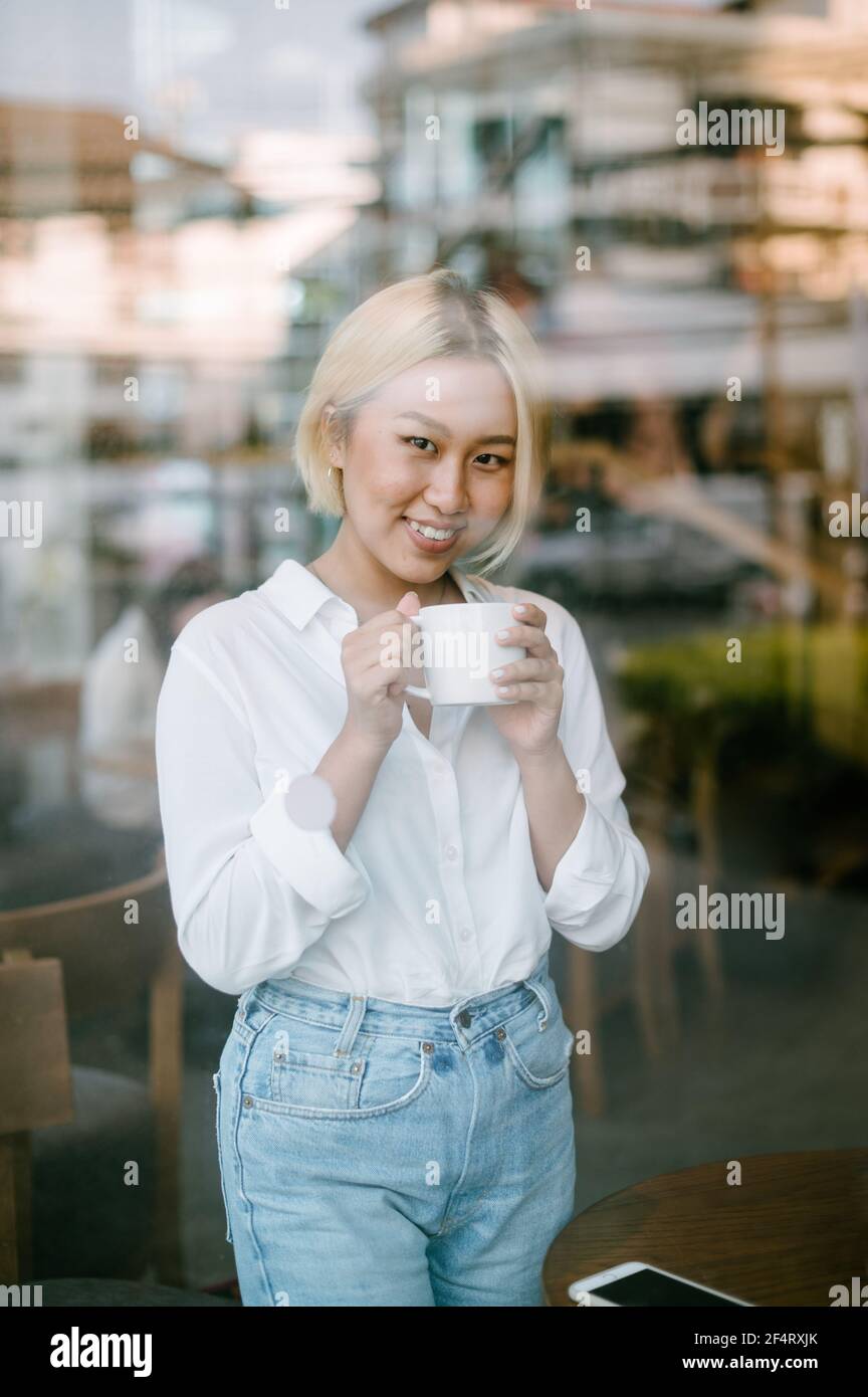 Une jeune femme asiatique avec une tasse de café dans ses mains est debout à l'intérieur derrière une grande fenêtre, souriant à l'extérieur de la caméra. Banque D'Images