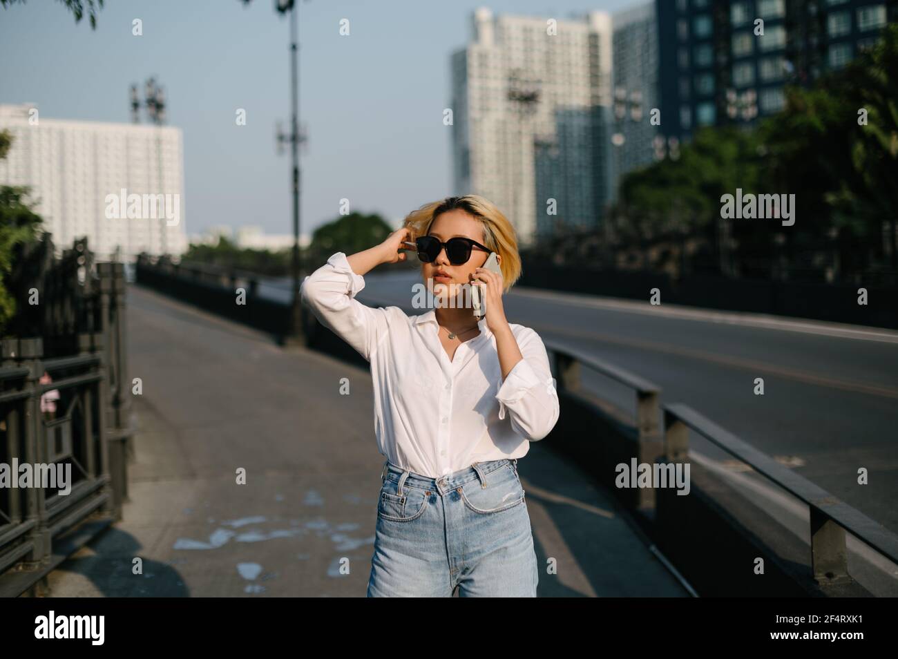 Une jeune femme asiatique avec des lunettes de soleil debout dans la rue parlant sur son téléphone, avec la route et les bâtiments à l'arrière-plan. Banque D'Images
