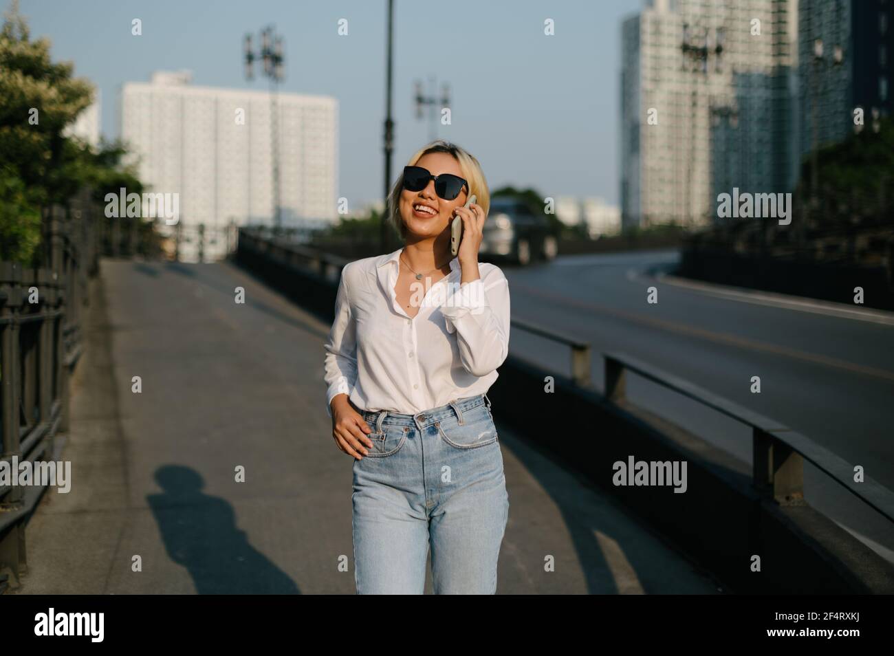 Une jeune femme asiatique avec des lunettes de soleil marchant dans la rue souriant joyeusement en parlant sur son téléphone. Une sensation de bien-être. Banque D'Images