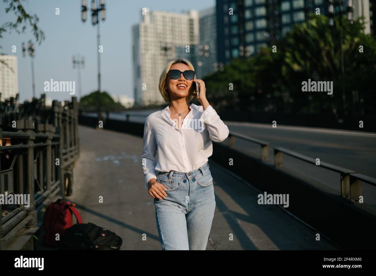 Une jeune femme asiatique avec des lunettes de soleil marchant dans la rue souriant joyeusement en parlant sur son téléphone. Une sensation de bien-être. Banque D'Images
