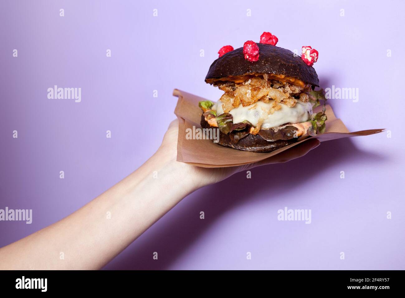 Cheeseburger Corona (antiCOVID) avec pain noir maison, laitue, oignon frit et fromage sur main féminine sur fond violet. Restaurant, Burger bar. Banque D'Images