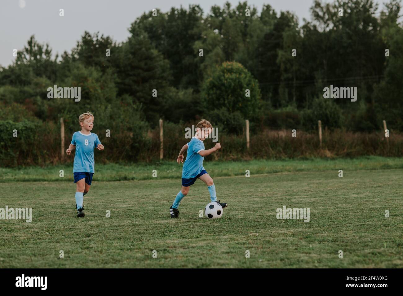 Enfants qui s'exécutent après le ballon de football. Longueur complète de garçons gais vêtues de robes de football bleues pourchassant le ballon sur le terrain de football. Banque D'Images