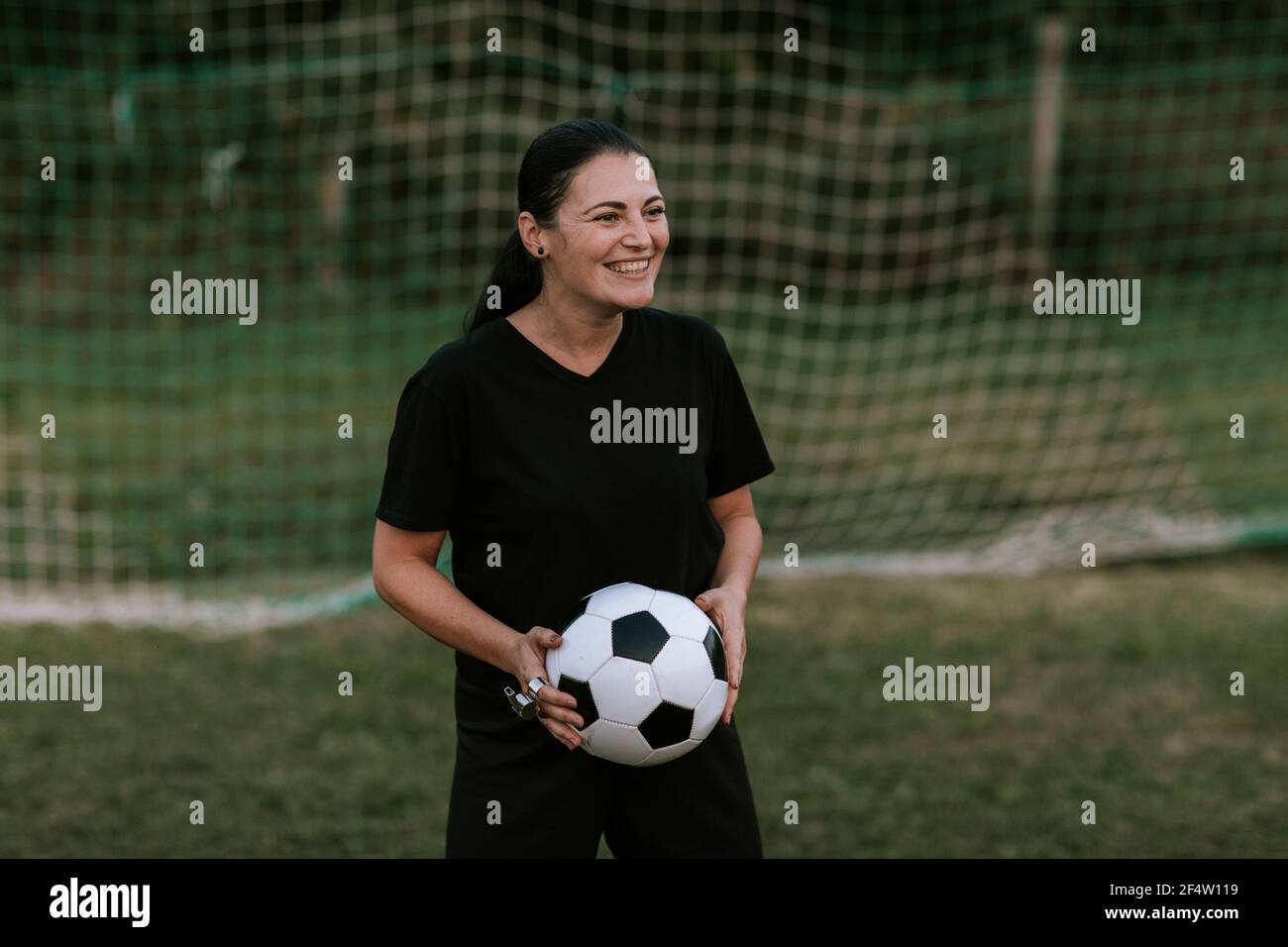 Une femme joyeuse arbitre de football se tenant au but sur le terrain de football. Femme amateur arbitre en robe de football noir tenant le ballon devant le filet de football. Banque D'Images