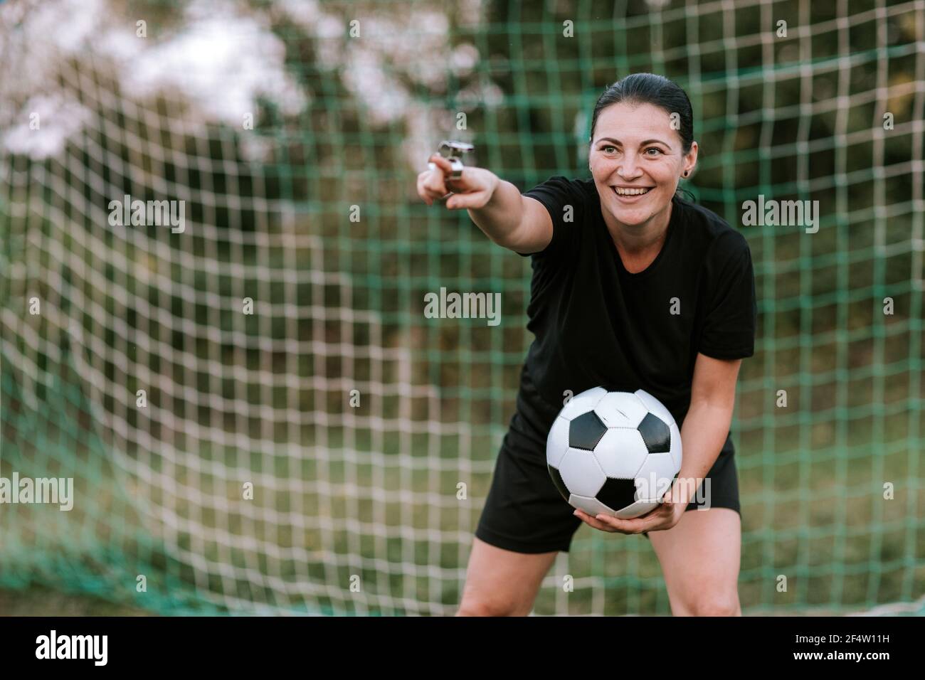 Une femme joyeuse arbitre de football se tenant au but sur le terrain de football. Femme amateur arbitre en robe de football noir tenant le ballon devant le filet de football. Banque D'Images