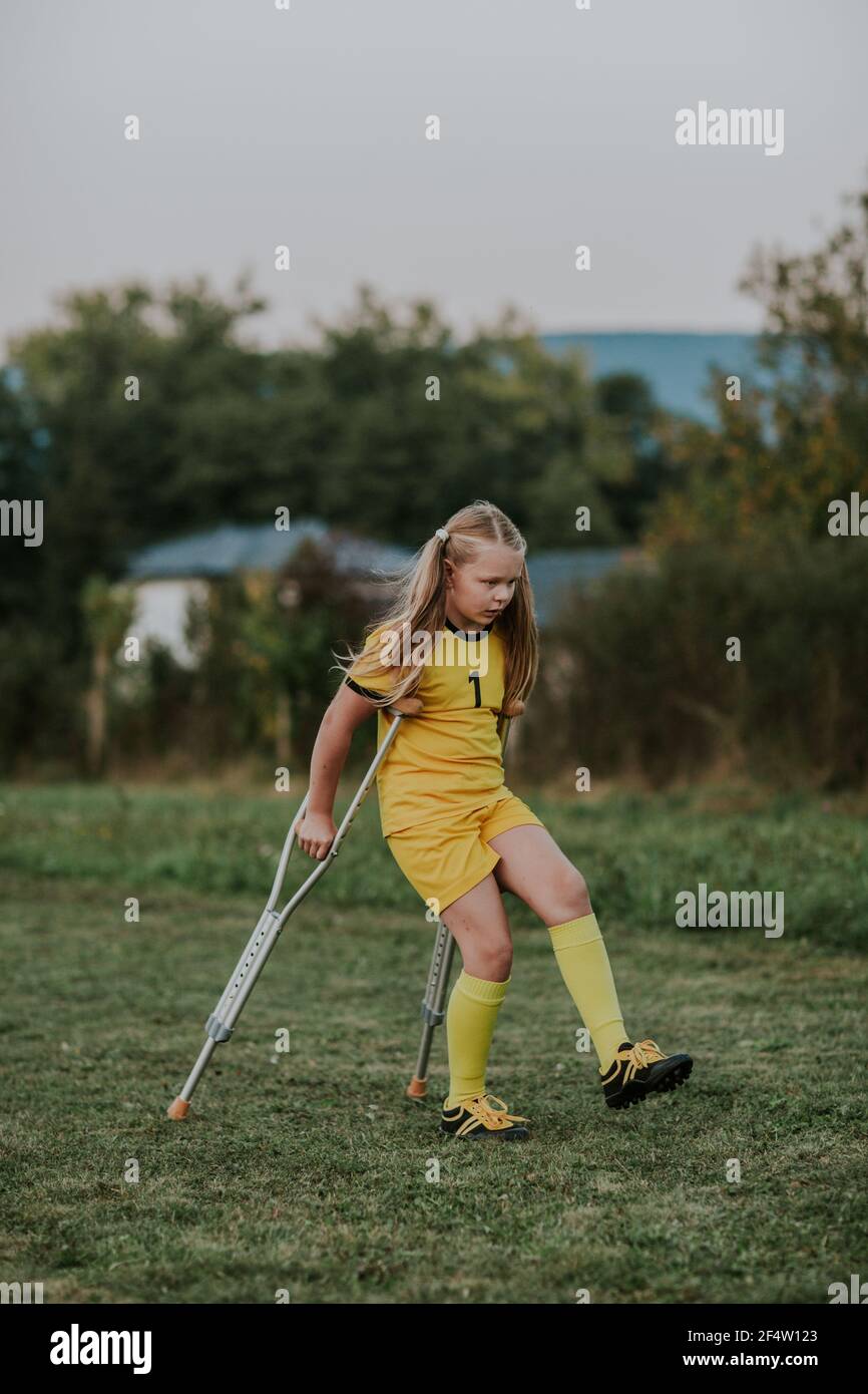 Fille avec des béquilles marchant le long du terrain de football. Pleine longueur de fillette blessée gardien de but dans la robe jaune de football sur les béquilles. Banque D'Images