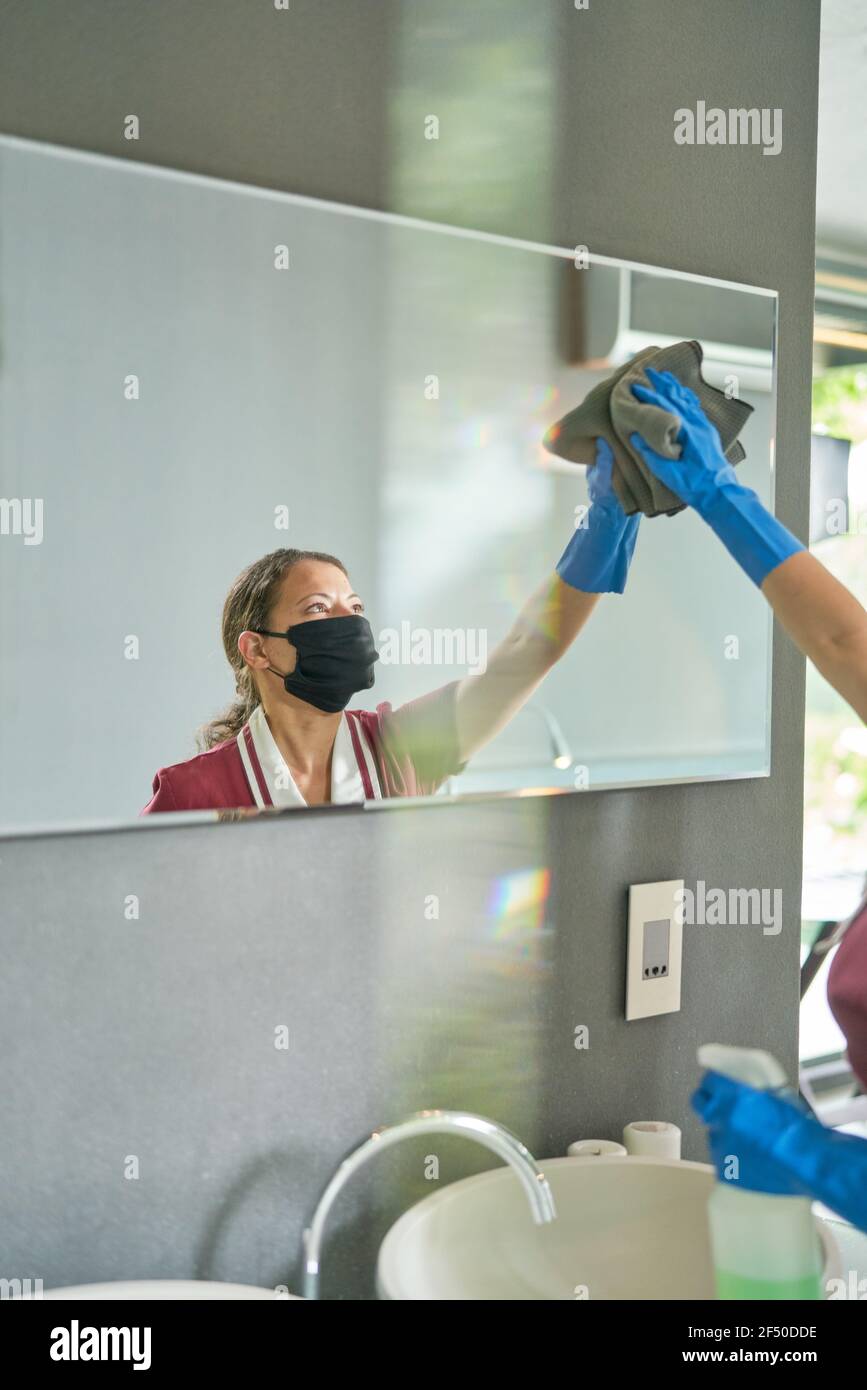 Femme de ménage dans un masque facial et des gants nettoyant le miroir dans la salle de bains Banque D'Images