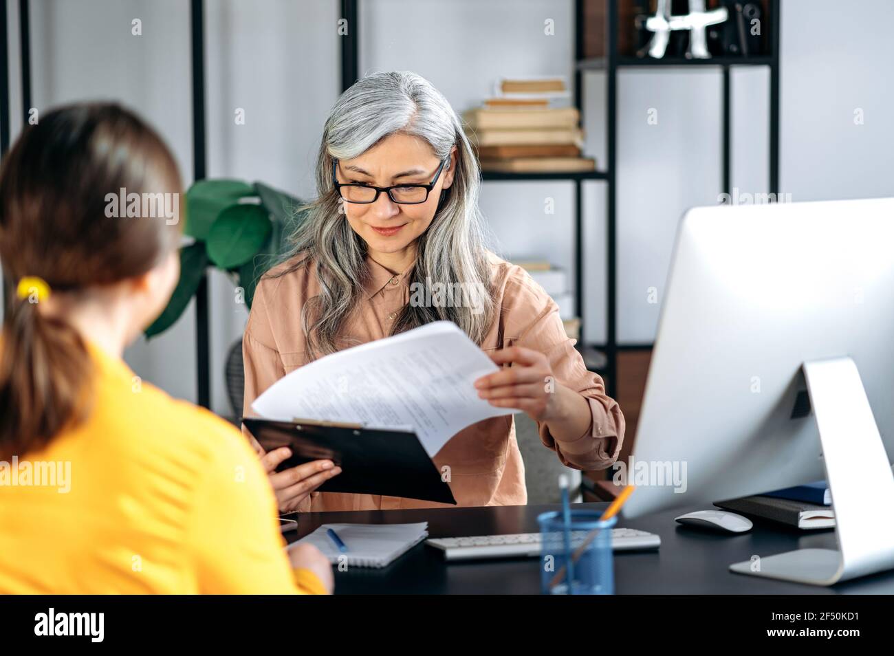 Intelligente confiante et influente femme asiatique aux cheveux gris portant des lunettes, femme d'affaires, responsable des ressources humaines, parlant à un candidat au poste dans un bureau moderne, tient et étudie à nouveau, souriant amical Banque D'Images