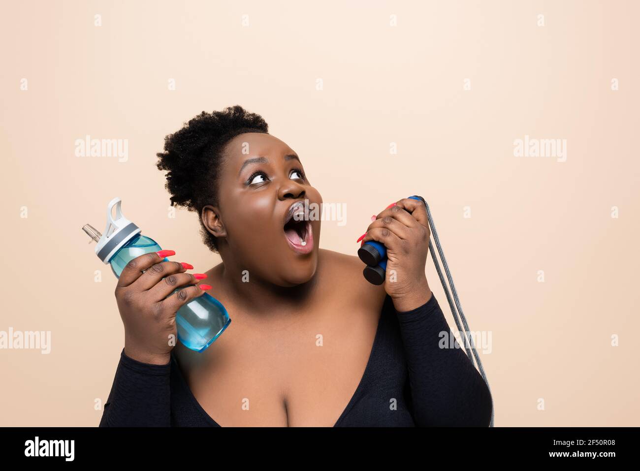 femme afro-américaine surdimensionnée dans des vêtements de sport tenant une bouteille de sport avec eau et corde à sauter isolée sur beige Banque D'Images