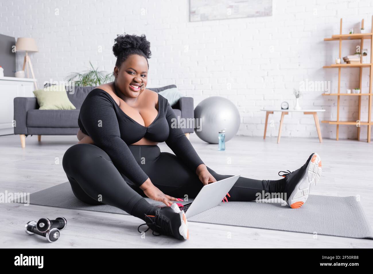 bonne femme afro-américaine de grande taille dans les vêtements de sport  assis sur tapis de fitness avec ordinateur portable Photo Stock - Alamy