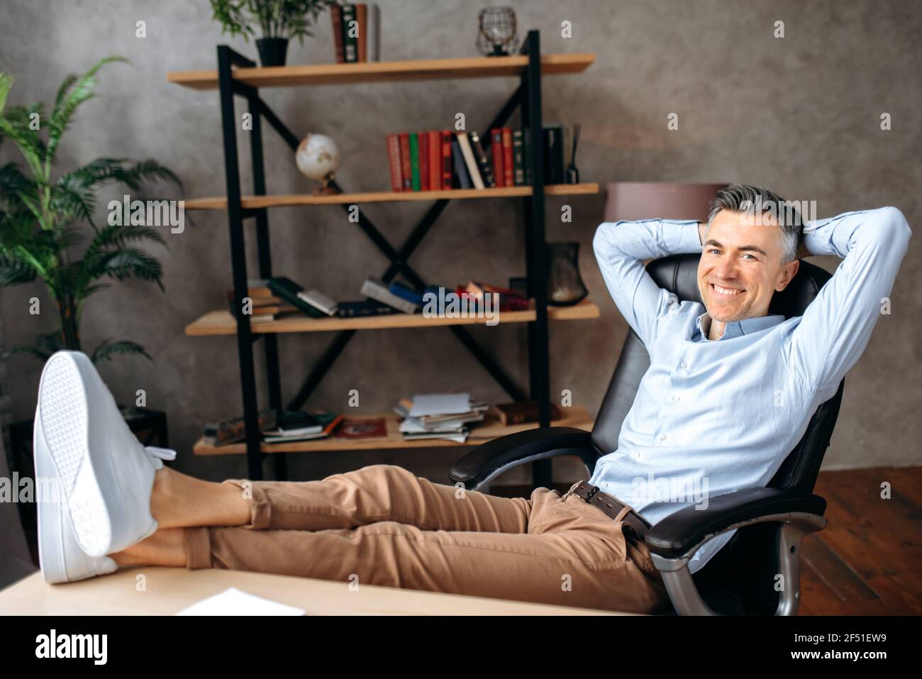 Beau homme d'affaires ou directeur caucasien mature, freelance se détend tout en s'asseyant avec ses jambes lancées sur son bureau, regardant la caméra, avec les mains derrière sa tête, souriant amical Banque D'Images