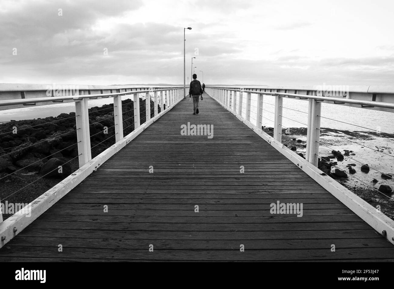 Monochrome noir et blanc d'un jeune homme marchant sur une jetée en bois au lever du soleil avec sa canne à pêche. Banque D'Images