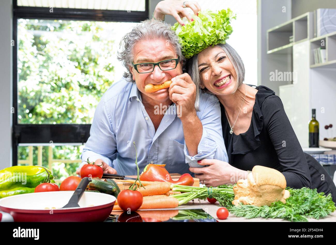 Couple senior s'amuser dans la cuisine avec des aliments sains - les gens fatigués cuisent repas à la maison avec l'homme et la femme préparant le déjeuner avec des légumes bio Banque D'Images