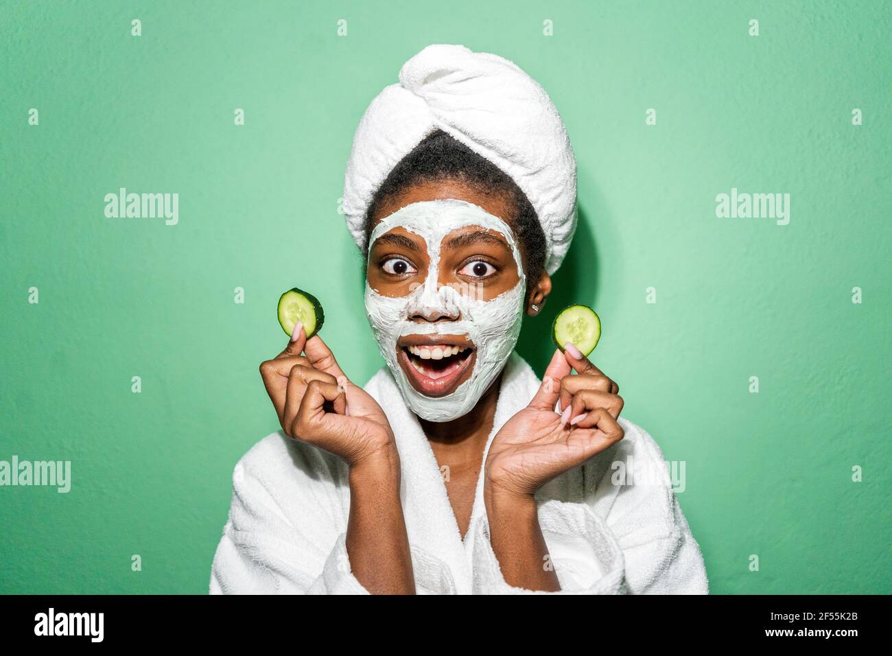 Jeune femme avec masque facial tenant le concombre tout en se tenant contre arrière-plan vert Banque D'Images