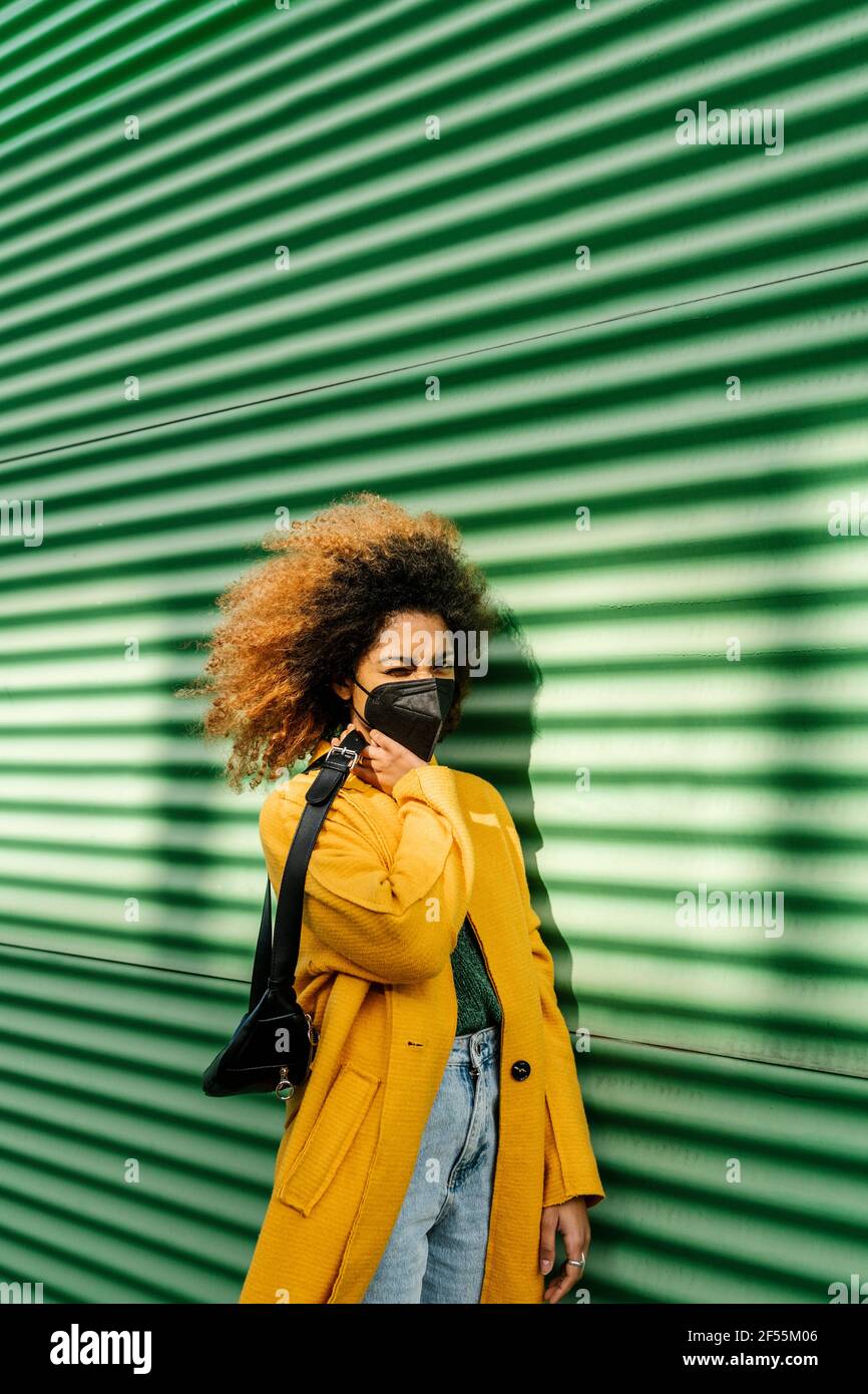 Femme afro avec masque de protection tenant la taille par Mur vert pendant COVID-19 Banque D'Images