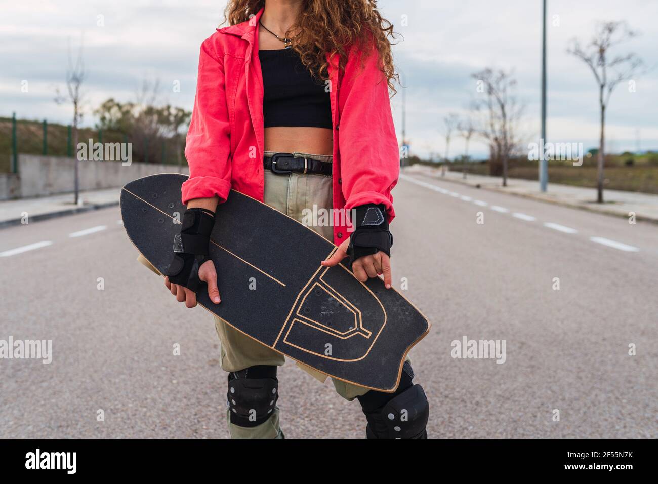 Femme portant des gants sans doigts qui fait des gestes tout en tenant le skateboard sur la route Banque D'Images