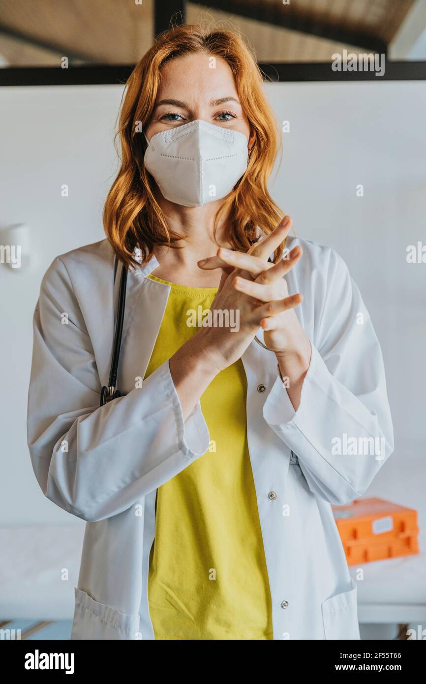 Femme médecin adulte de taille moyenne portant un masque facial désinfectant les mains avec assainisseur en se tenant à la clinique Banque D'Images