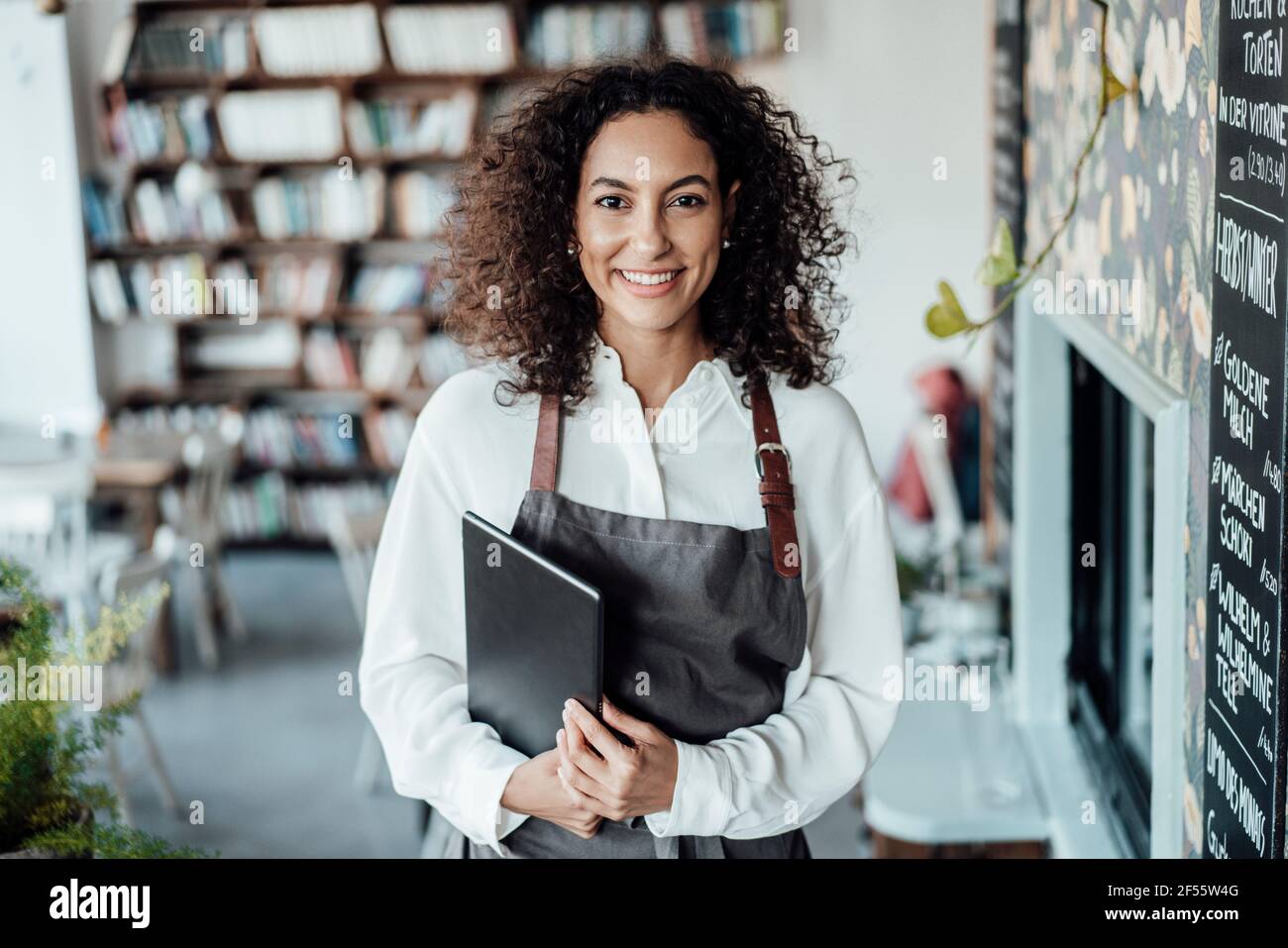 Femme souriante portant un tablier tenant un livre tout en se tenant au café Banque D'Images