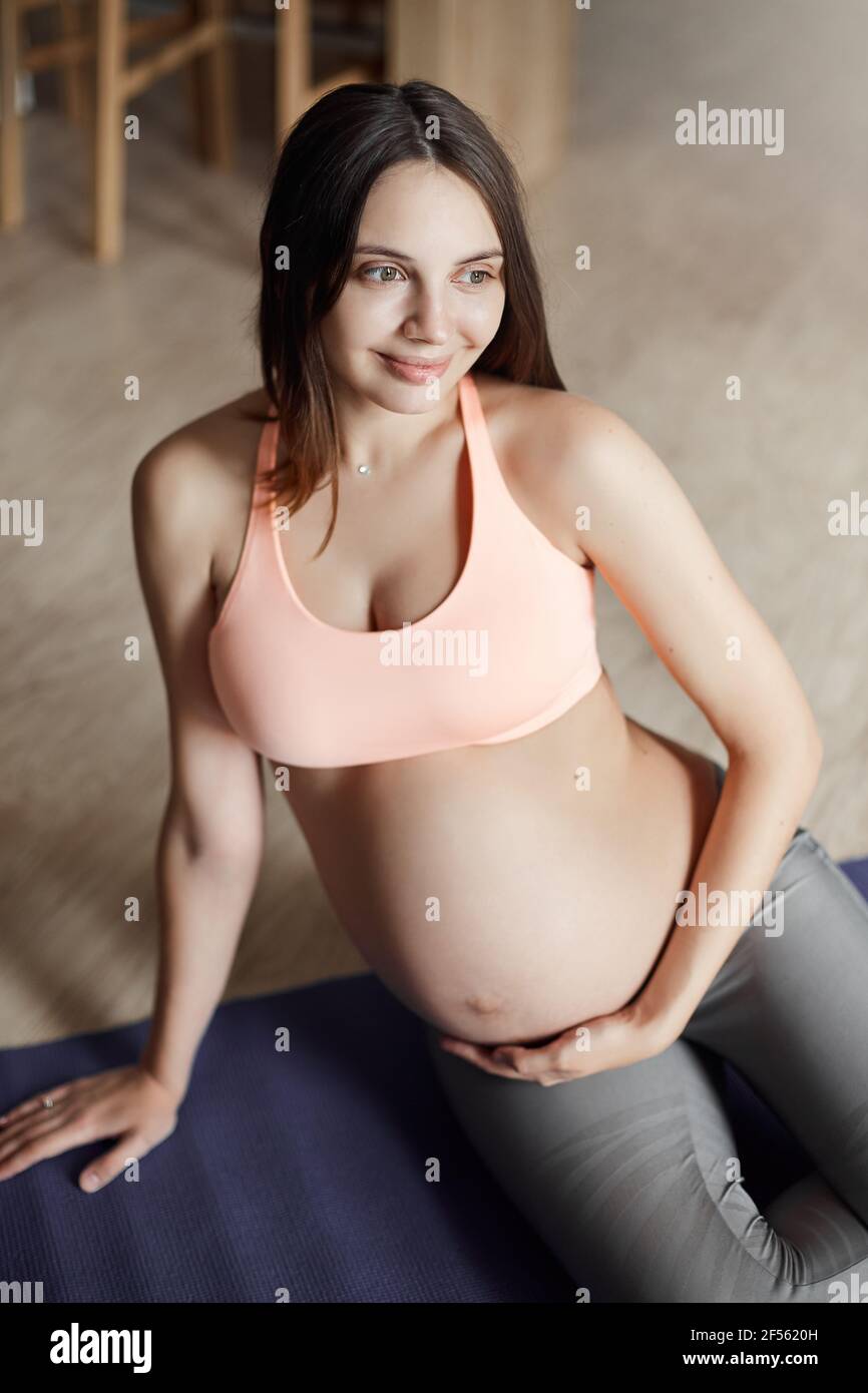 Photo verticale de la jeune femme enceinte européenne mignonne et heureuse  dans un soutien-gorge de sport et des leggings, se penchant à la main tout  en étant allongé sur un coussin, en