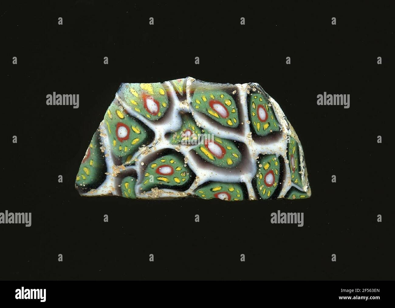 Fragment d'un vaisseau. Une faible courbure du fragment de verre permet l'affectation à un vaisseau, éventuellement à une coque. Dans une grille en nid d'abeille de lignes blanches, une fleur en technologie millefiori est toujours reconnaissable. Pour reconnaître un noyau blanc avec un contour rouge, des points jaunes sont visibles dans une bande circonférentielle verte. L'espace entre la fleur et le nid d'abeilles blanc est violet.pour la production différents verres de couleur aux tubes et tiges ont été façonnés. Elles étaient comprimées et chauffées, ce qui entraînait une longue barre. Le verre coupé en tranches de ​​now avait des motifs amorphes ou floraux différents. Par la suite, plusieurs de ces platele Banque D'Images